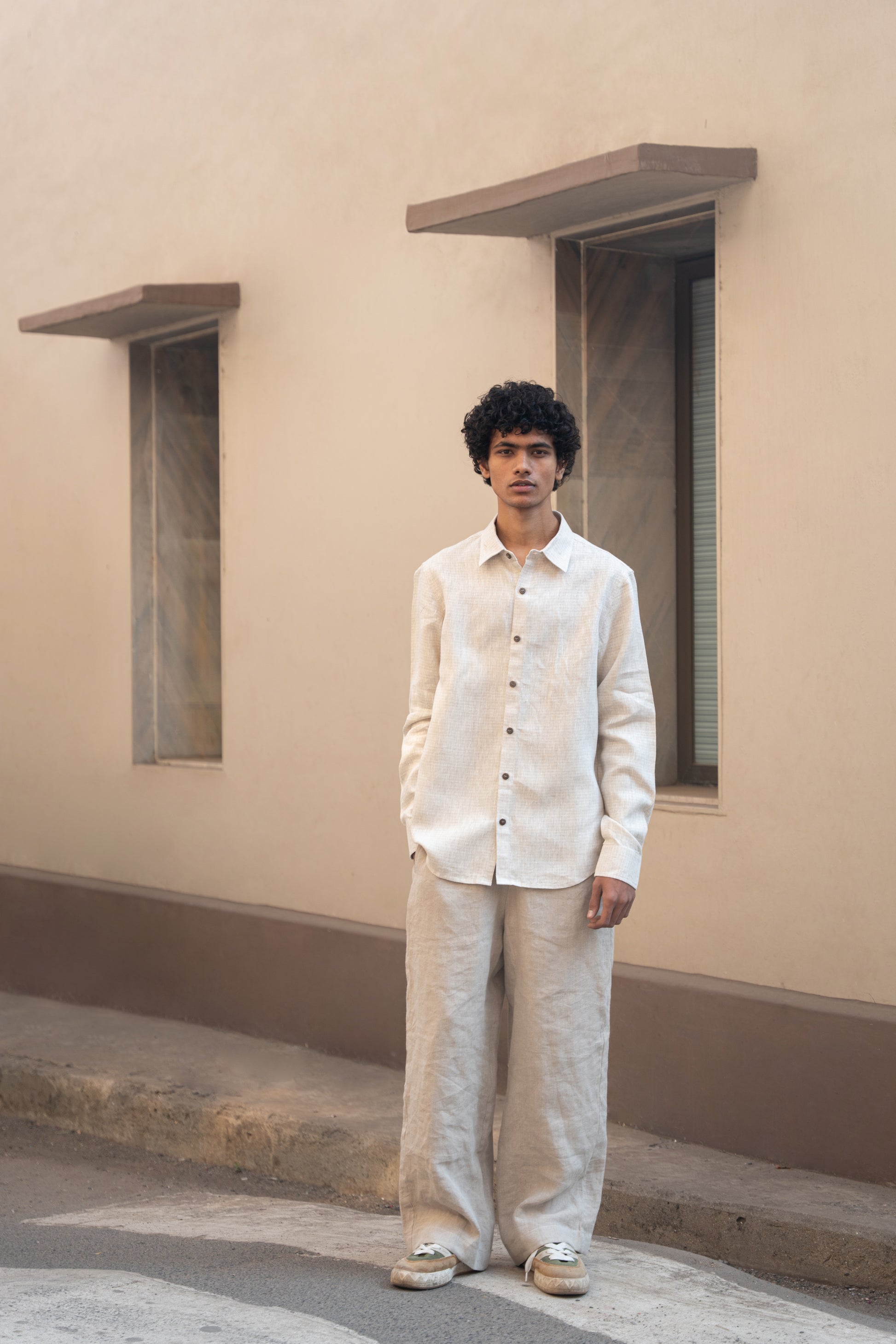 A man in light beige full sleeved pure linen shirt and a darker beige pants is standing in front  of a wall