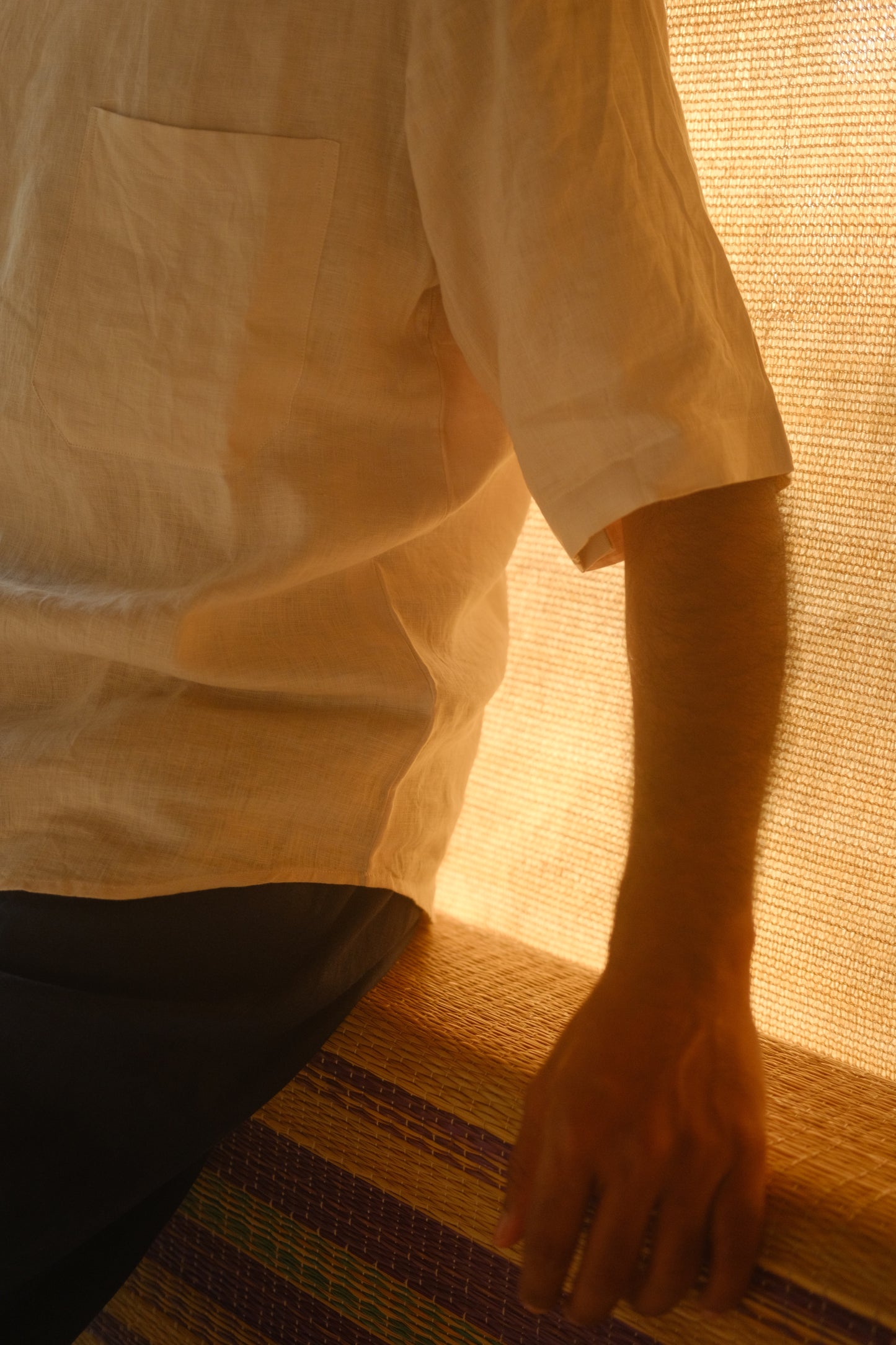 Close up of the pocket detail of an off-white linen shirt worn by a man sitting on a bamboo rug