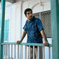 A young man in a blue linen half-sleeved shirt with hand-embroidered kantha motives of the hunter and gatherers life is standing on a window railing