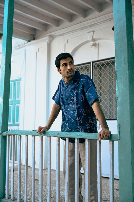 A young man in a blue linen half-sleeved shirt with hand-embroidered kantha motives of the hunter and gatherers life is standing on a window railing