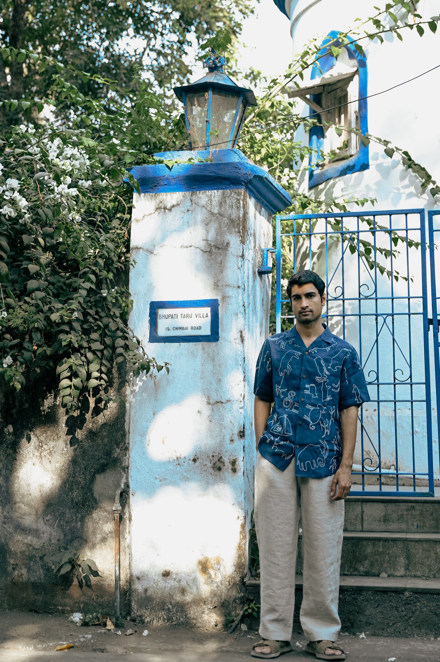 A young man in a blue linen half-sleeved shirt with hand-embroidered kantha motives of the hunter and gatherers life is standing in front of a beautiful house