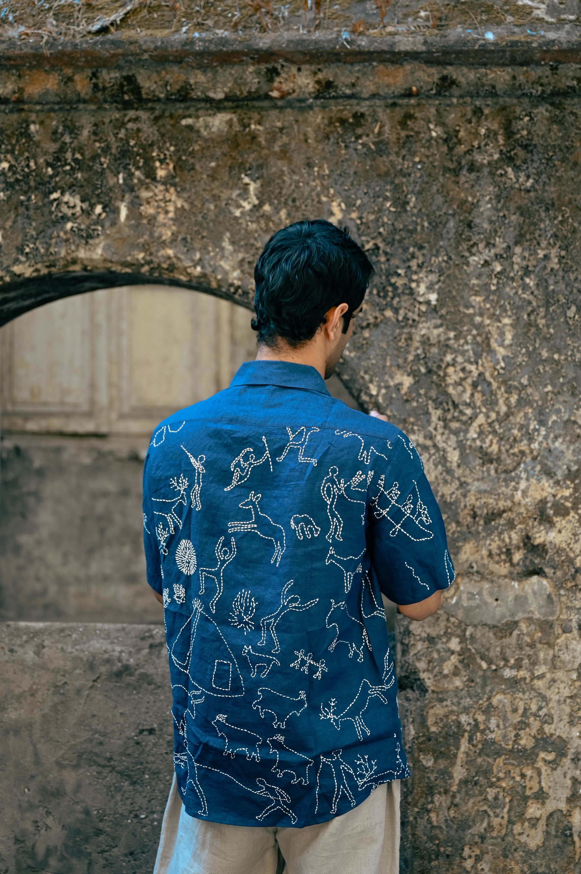 Back of a young man in a blue linen half-sleeved shirt with hand-embroidered kantha motives of the hunter and gatherers life is standing in front of a rustic wall