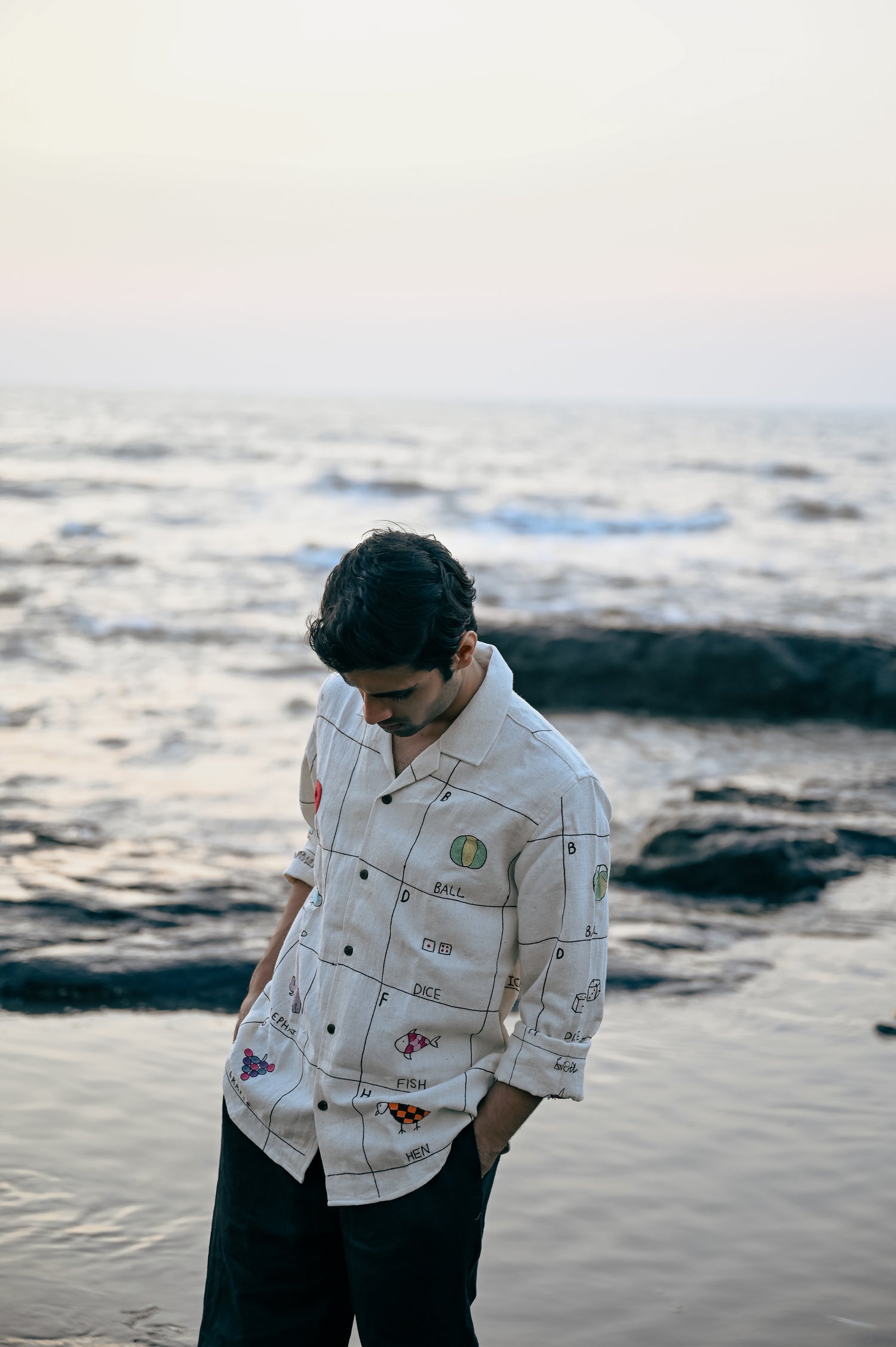 A young man in an beige linen full-sleeved shirt with the hand-embroidered alphabet is standing on the beach