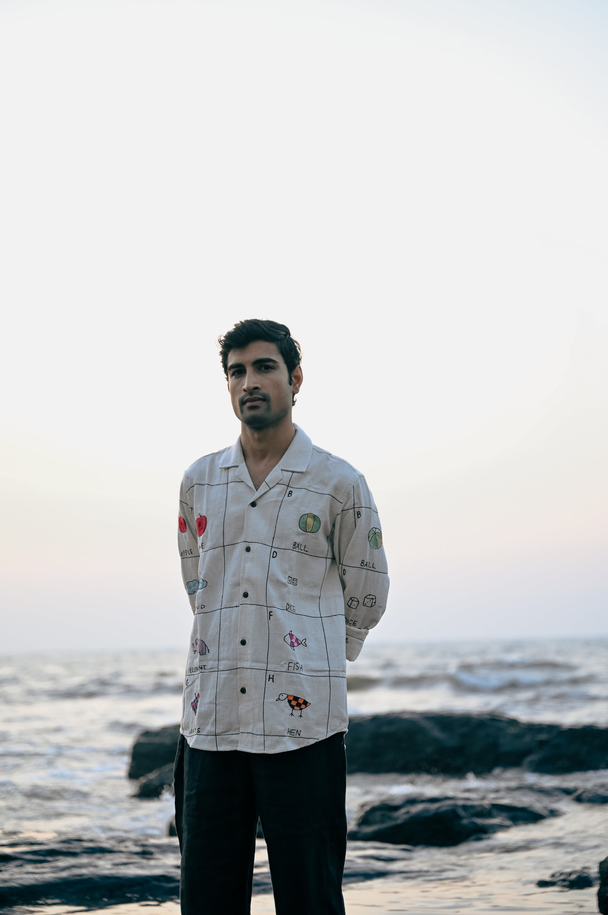 A young man in an beige linen full-sleeved shirt with the hand-embroidered alphabet is standing on the beach