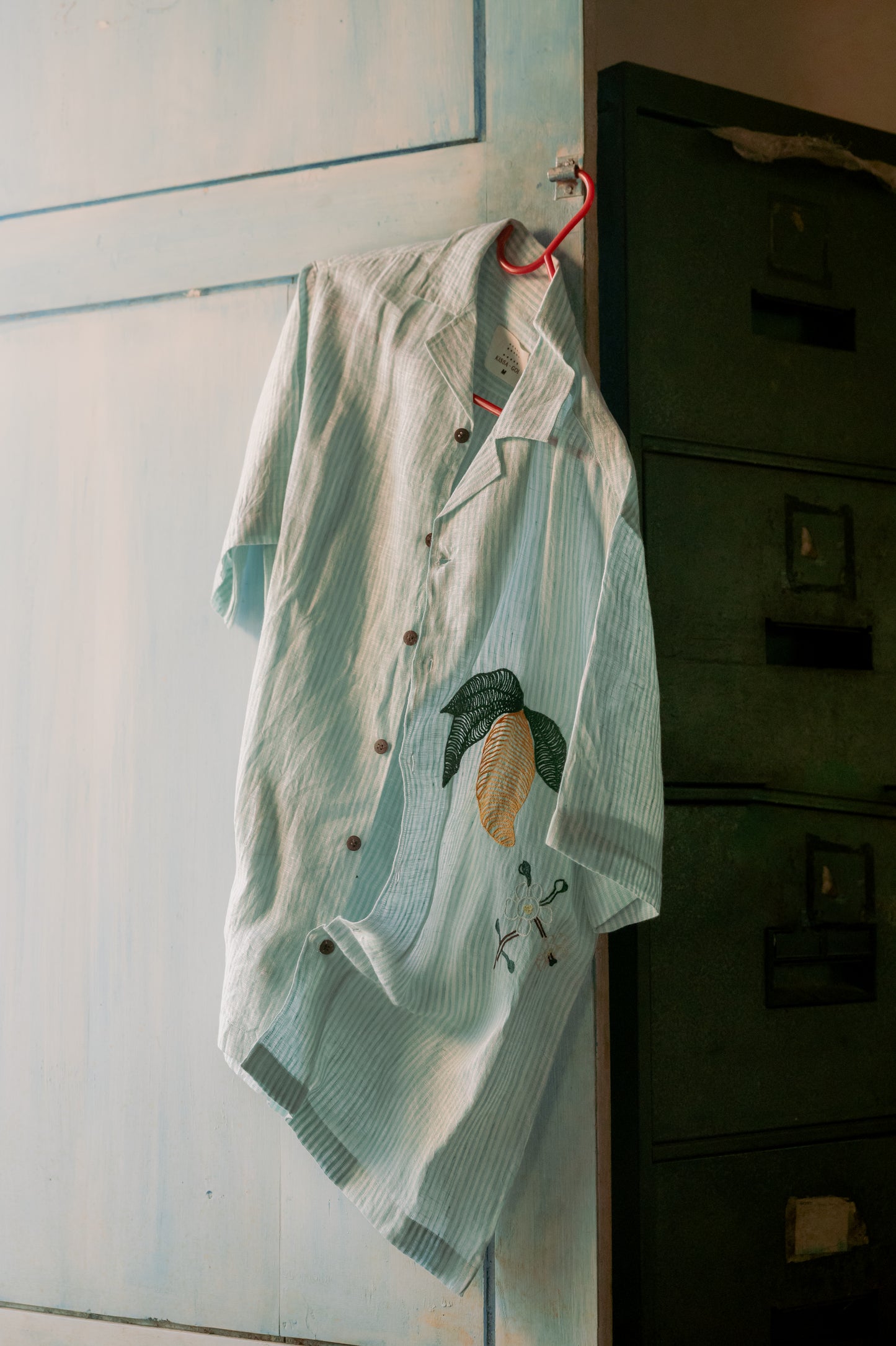 Green and white stripes linen shirt with embroidered mangoes hung on a blue door.