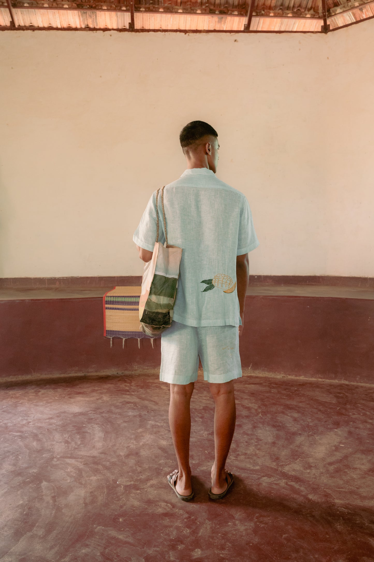 Back view of a man standing in a yoga hall wearing green and white stripes linen half-sleeved shirt and shorts with hand- embroidered mangoes.