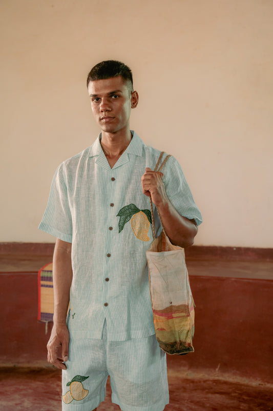 a man standing in a yoga hall wearing co-ordinated green and white stripe linen shirt and shorts with embroidered mangoes. Front view