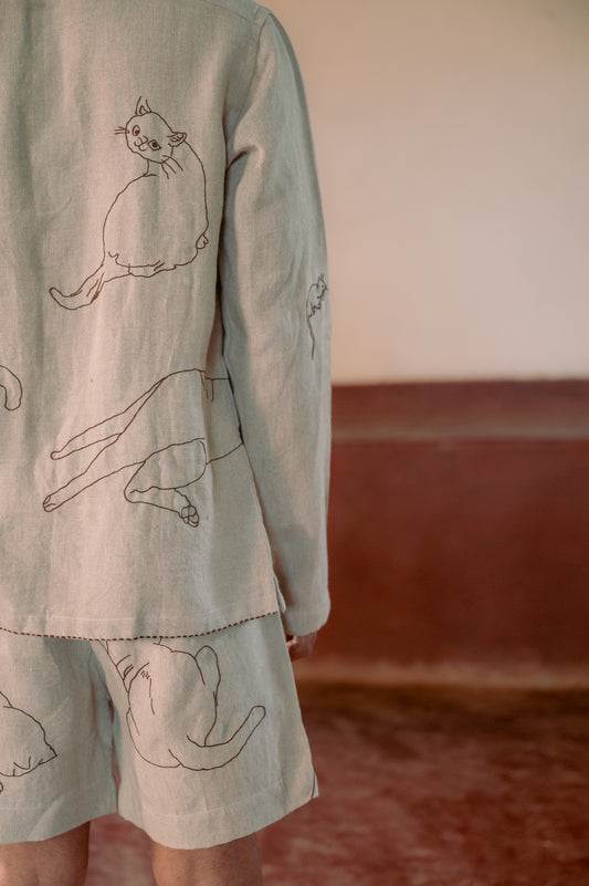 a right side Back view of a man standing in a yoga hall wearing beige linen full-sleeved shirt and shorts with hand- embroidered cats.