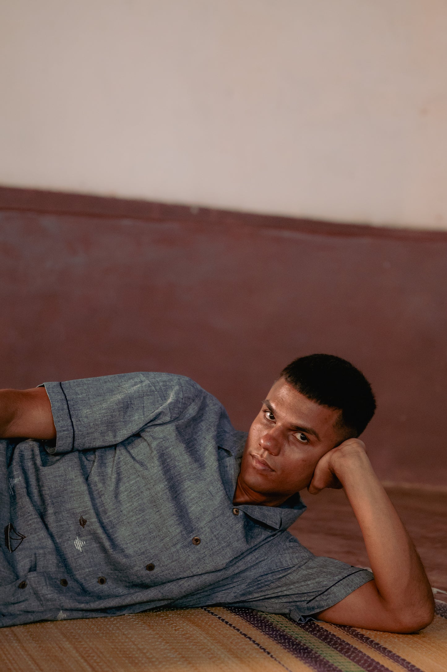 A man in blue organic cotton half-sleeved shirt with boats embroidered on the lower half of the shirt is lying on a yoga mat with his head propped up on his hand