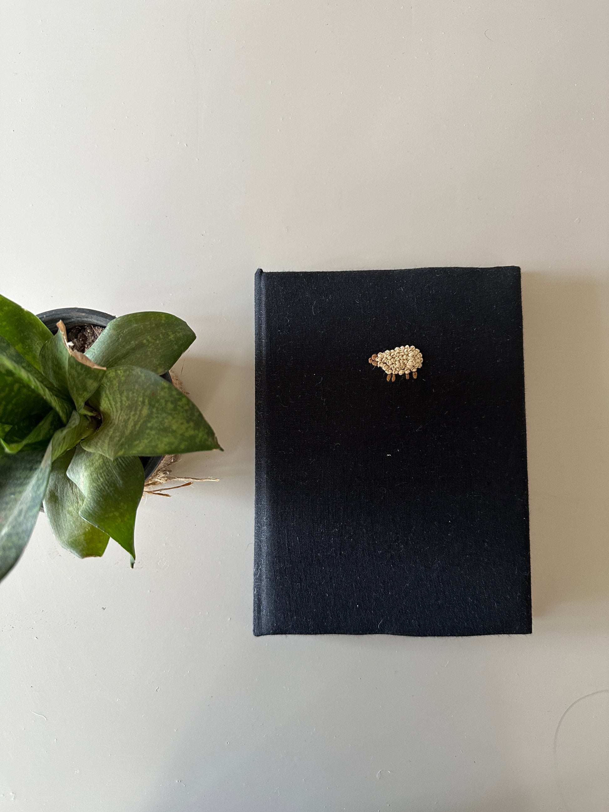 A black cover diary made with black linen scraps with a beige sheep embroidered on the cover placed next to a plant