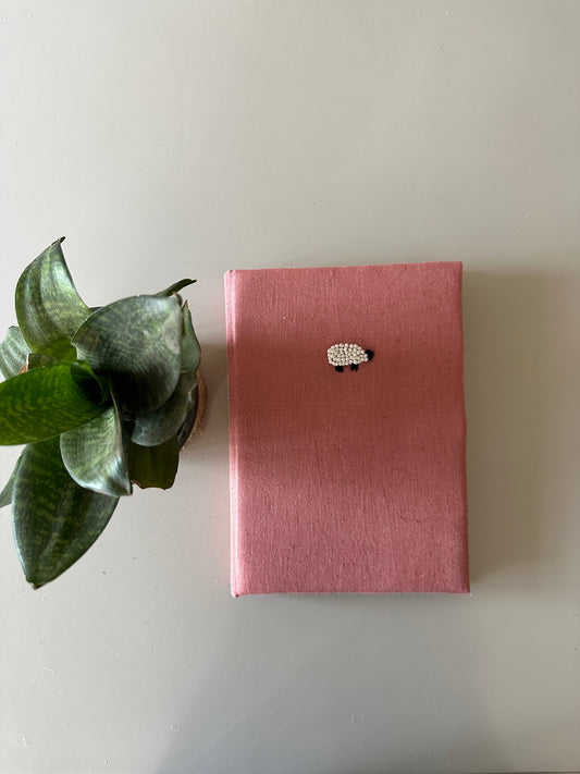 A peach cover diary made with peach linen scraps with a white sheep embroidered on the cover placed next to a plant