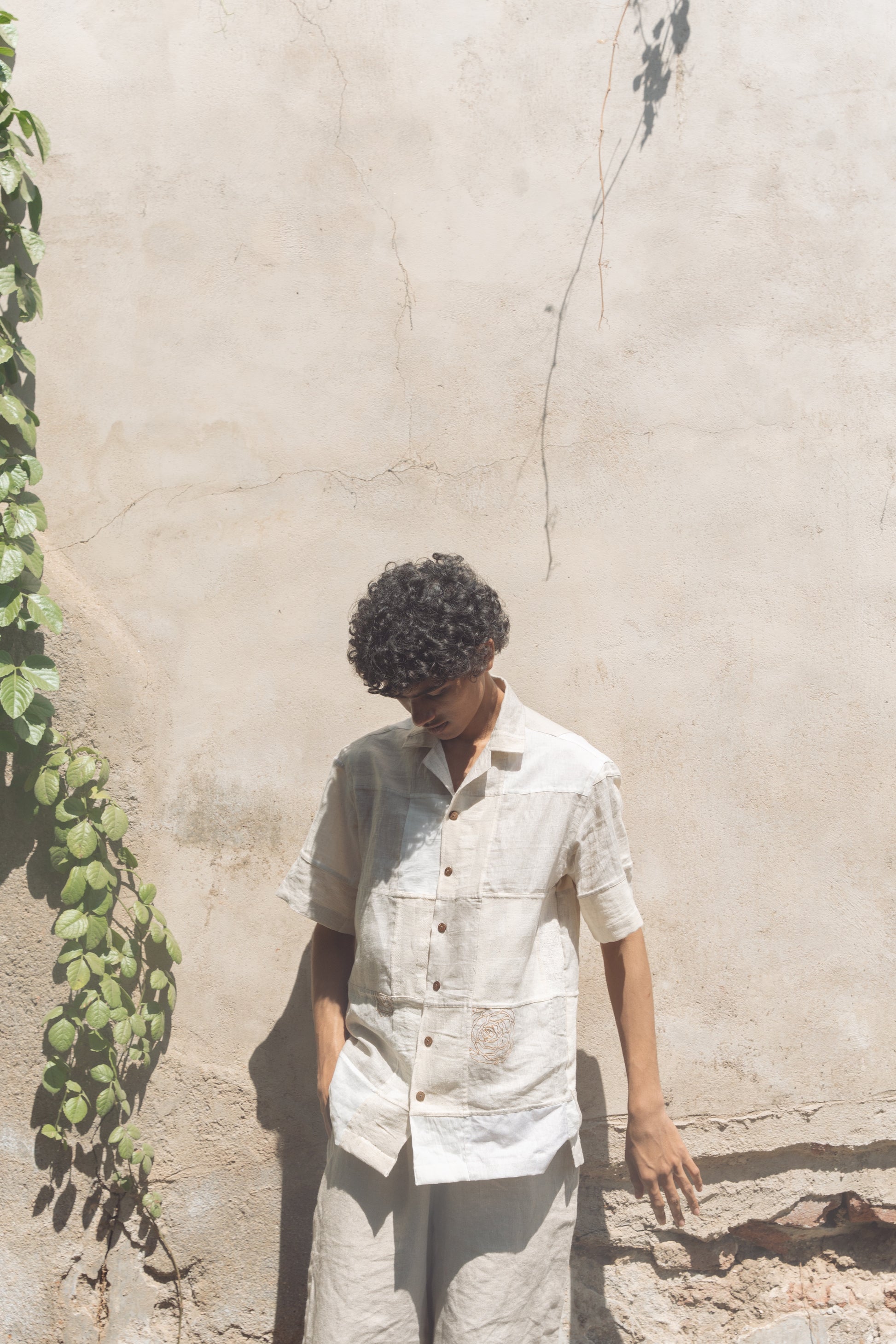 A young man in an off-white half-sleeved shirt made with patches of cotton and linen squares is standing in front of a white wall