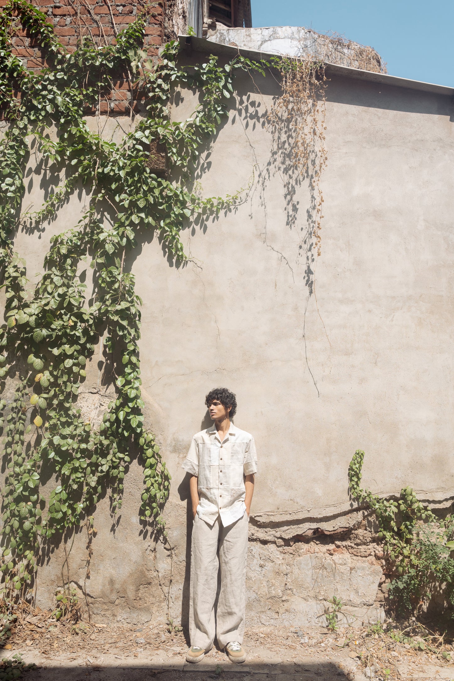 A young man in an off-white half-sleeved shirt made with patches of cotton and linen squares is standing in front of a white wall