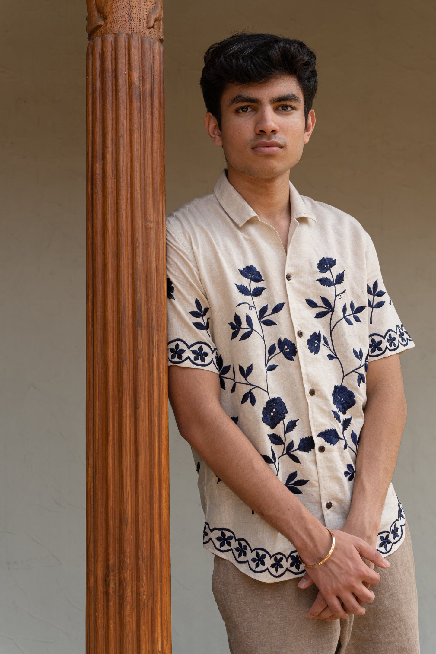 a man wearing beige linen blue floral hand-embroidered shirt leaning against a pillar.