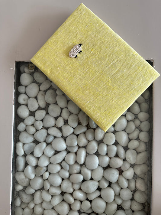 A yellow diary made with lemon yellow linen scraps with a white sheep embroidered on the cover lying on top of white stones