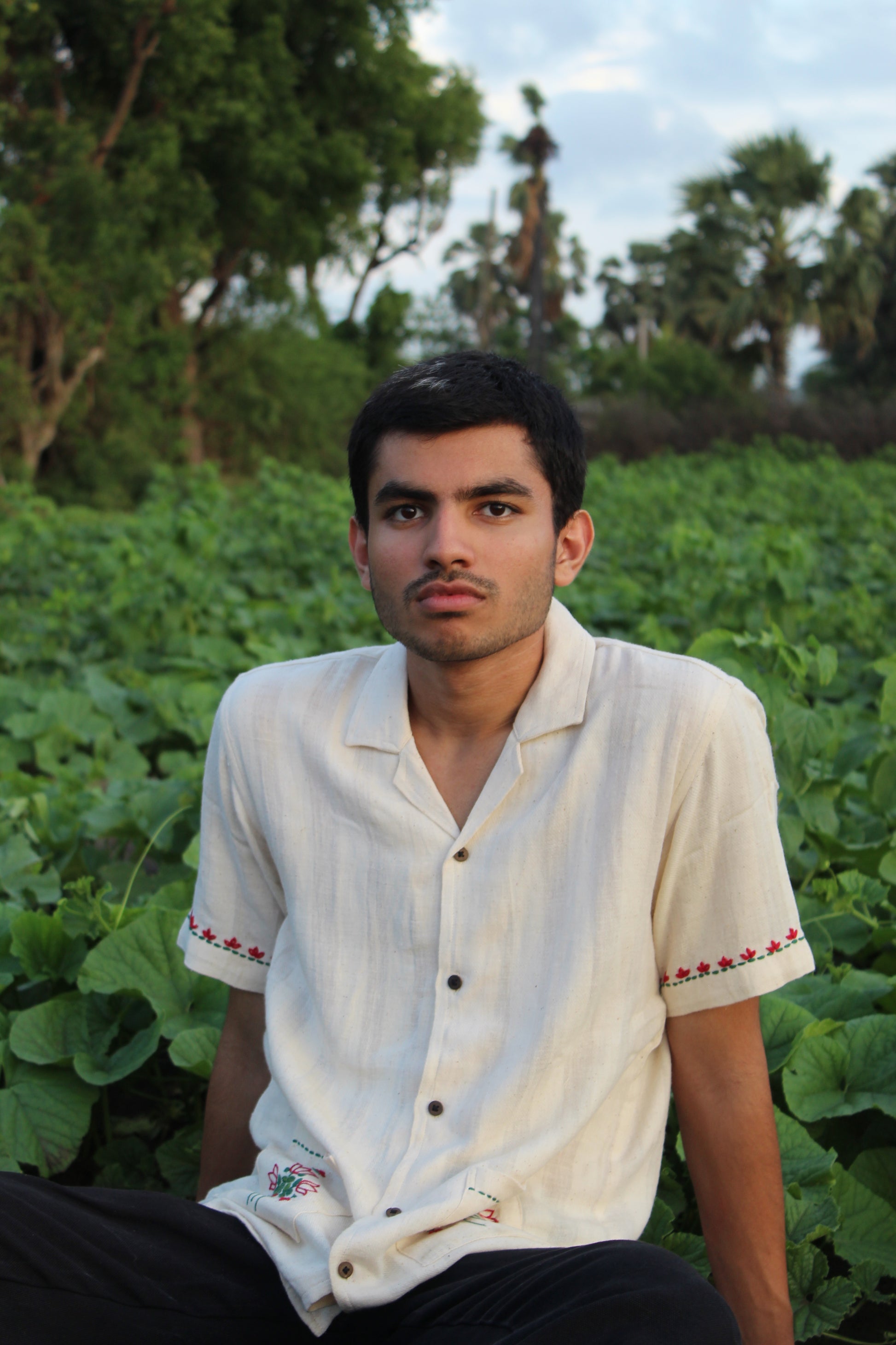 A young man in an off-white cotton shirt with emmbroidered pockets sitting in the middle of fields