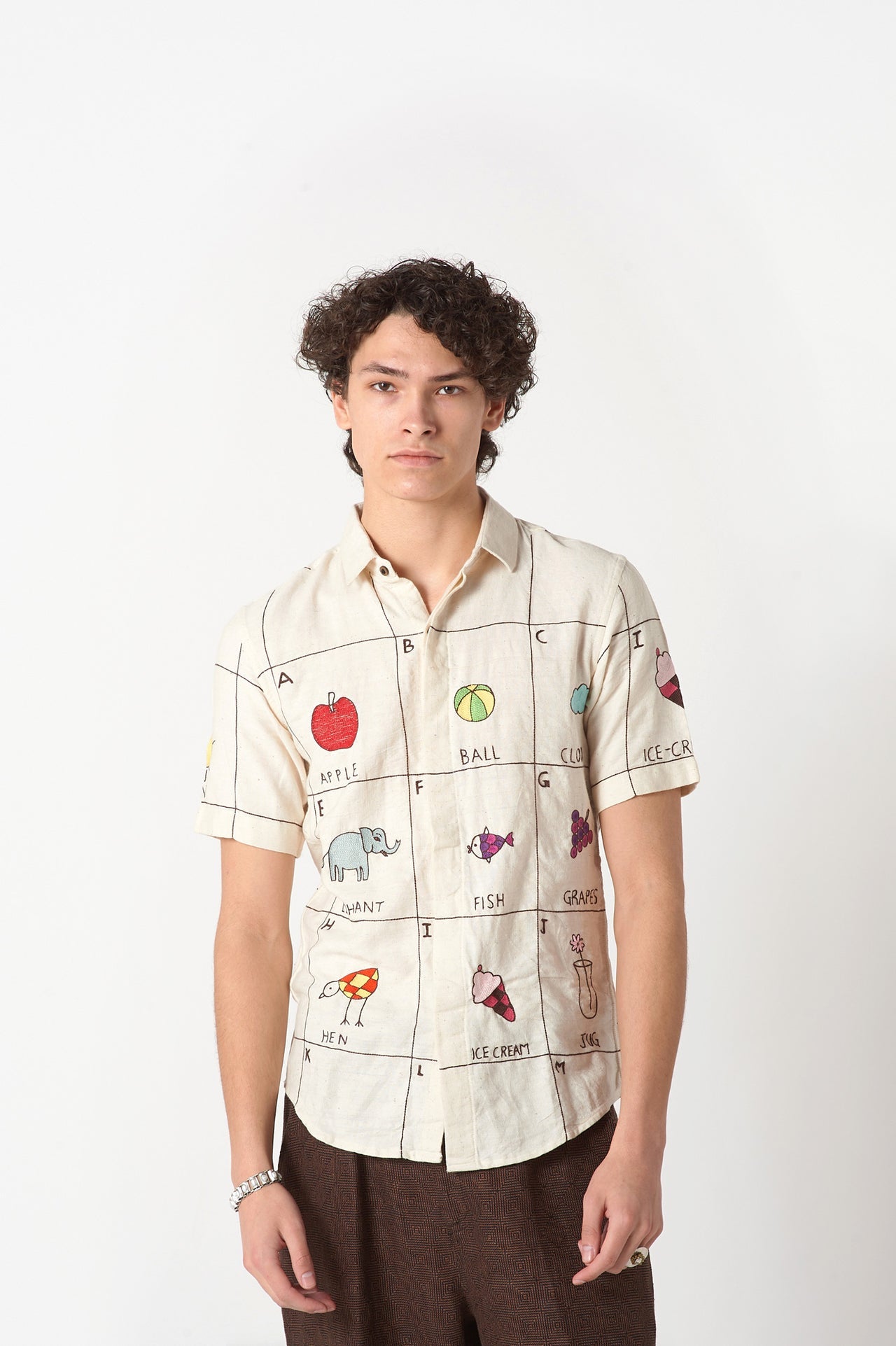 A young man in an offwhite cotton shirt with the hand-embroidered alphabet standing against a white wall