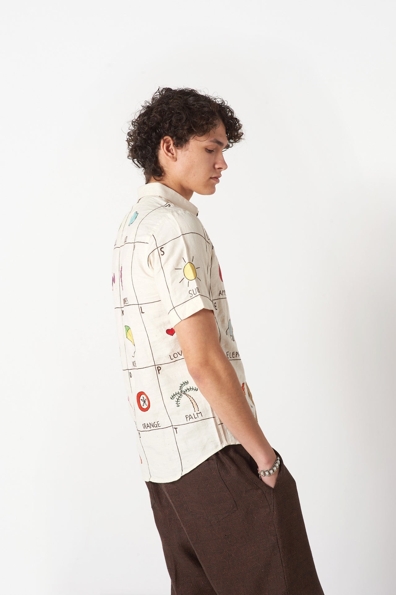 Side view of a young man in an offwhite cotton shirt with the hand-embroidered alphabet standing against a white wall