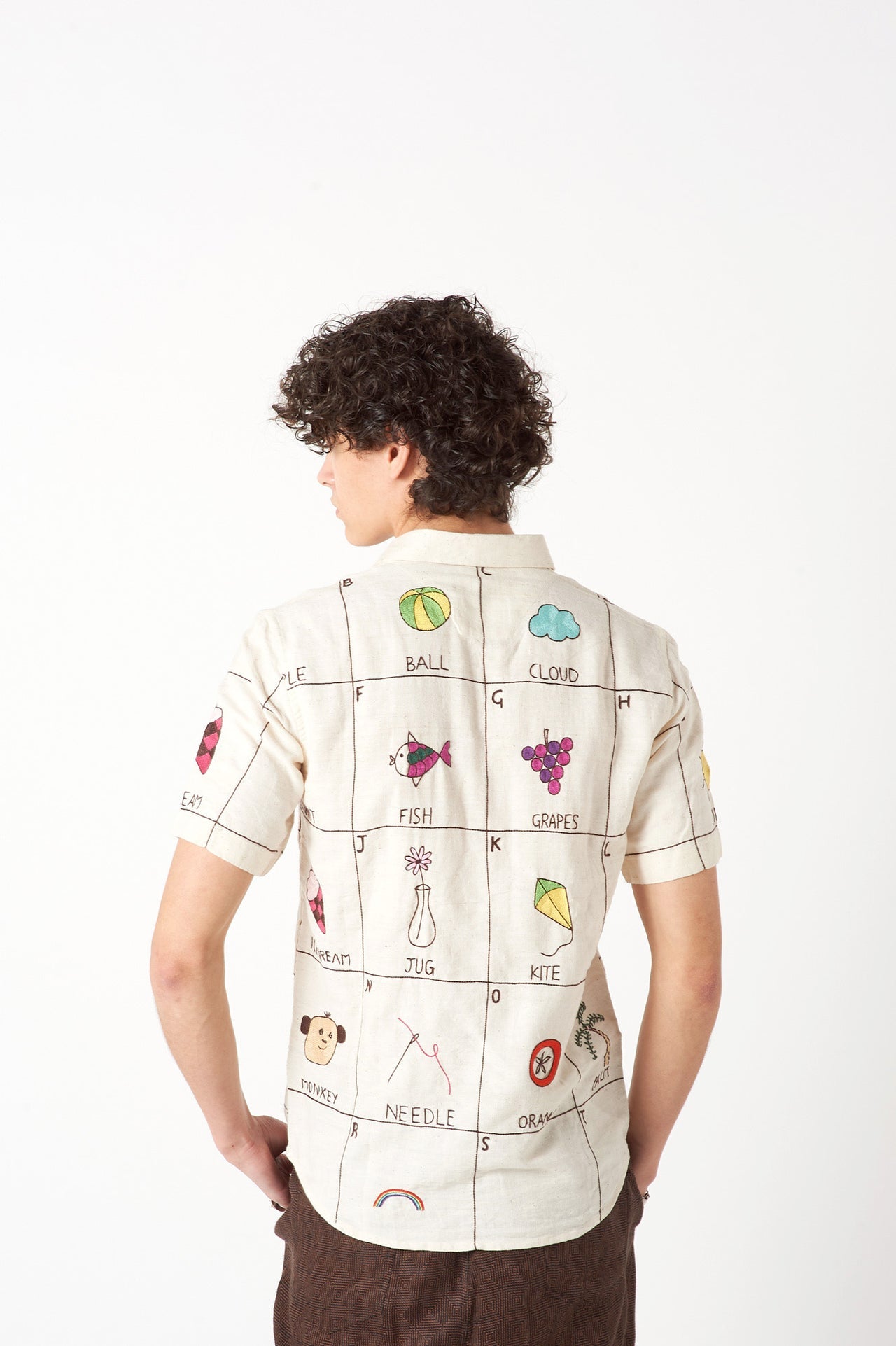 Back detail of a young man in an offwhite cotton shirt with the hand-embroidered alphabet standing against a white wall