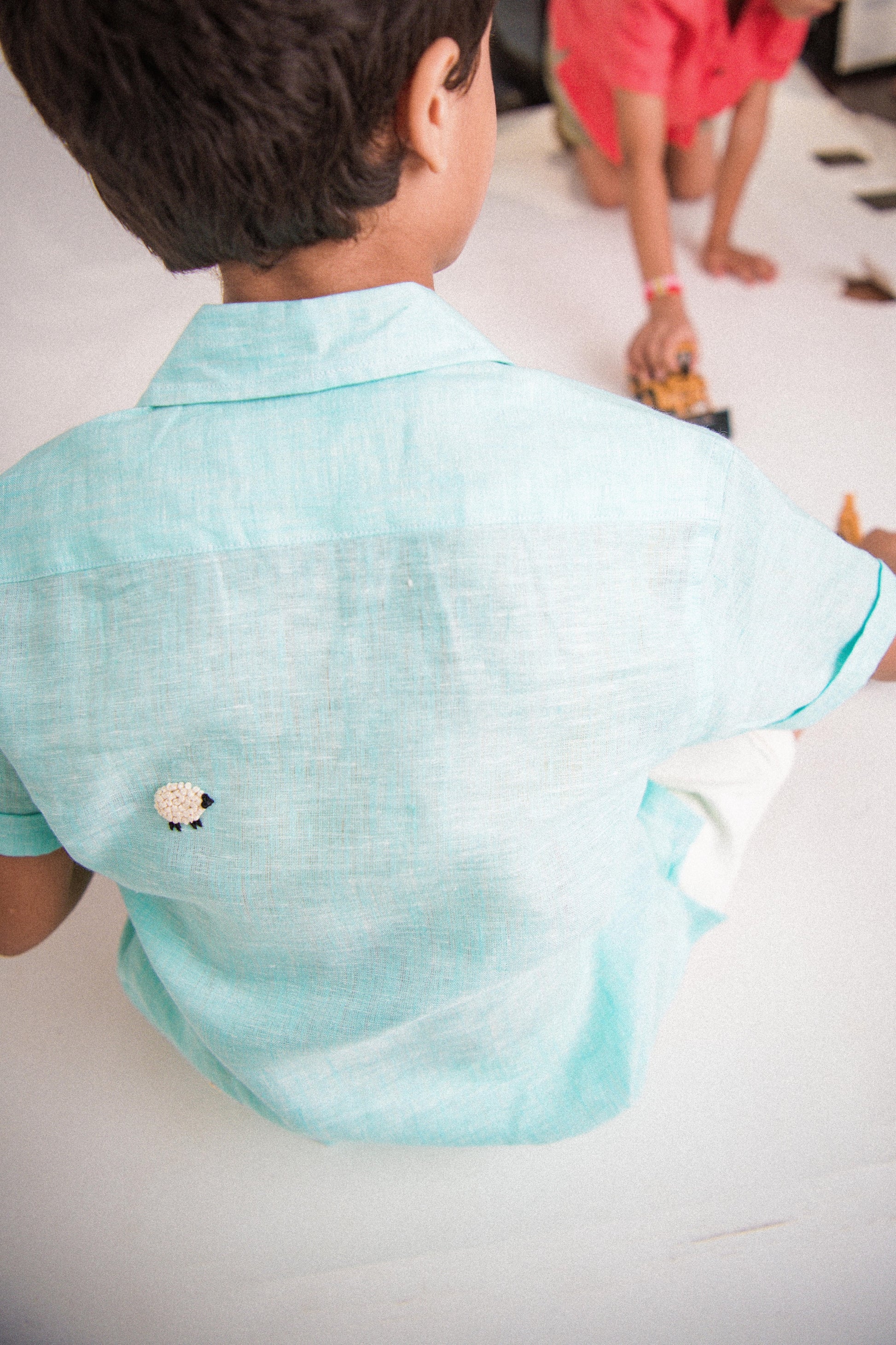 Back details of a young 7-8 year old boy in a blue linen half sleeved shirt with small sheep embroidered throughout is sitting against a white background