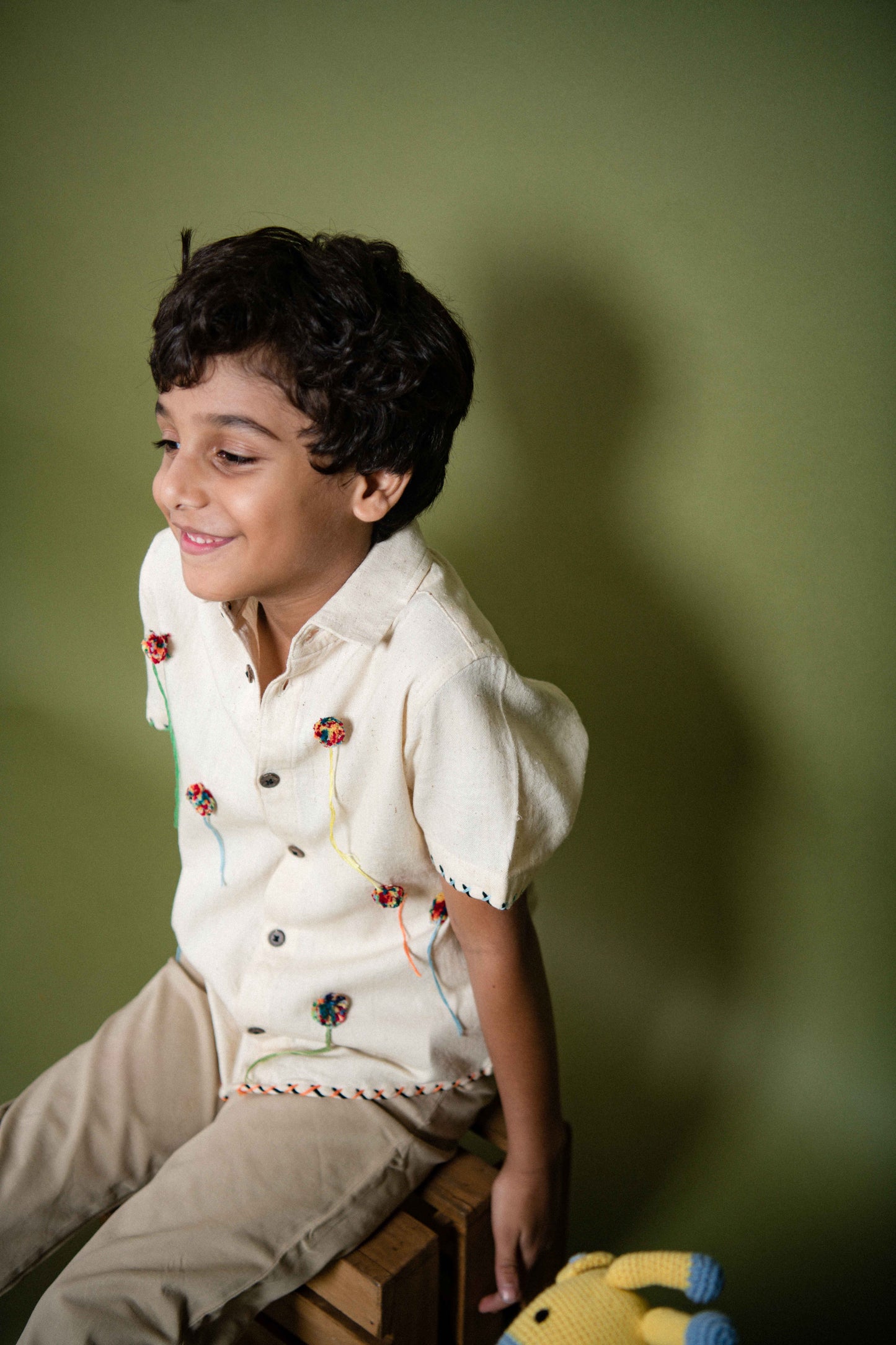 Side view of a 6 year old kid in our kala cotton half-sleeved shirt with pom poms embroidered through out sitting on a crate