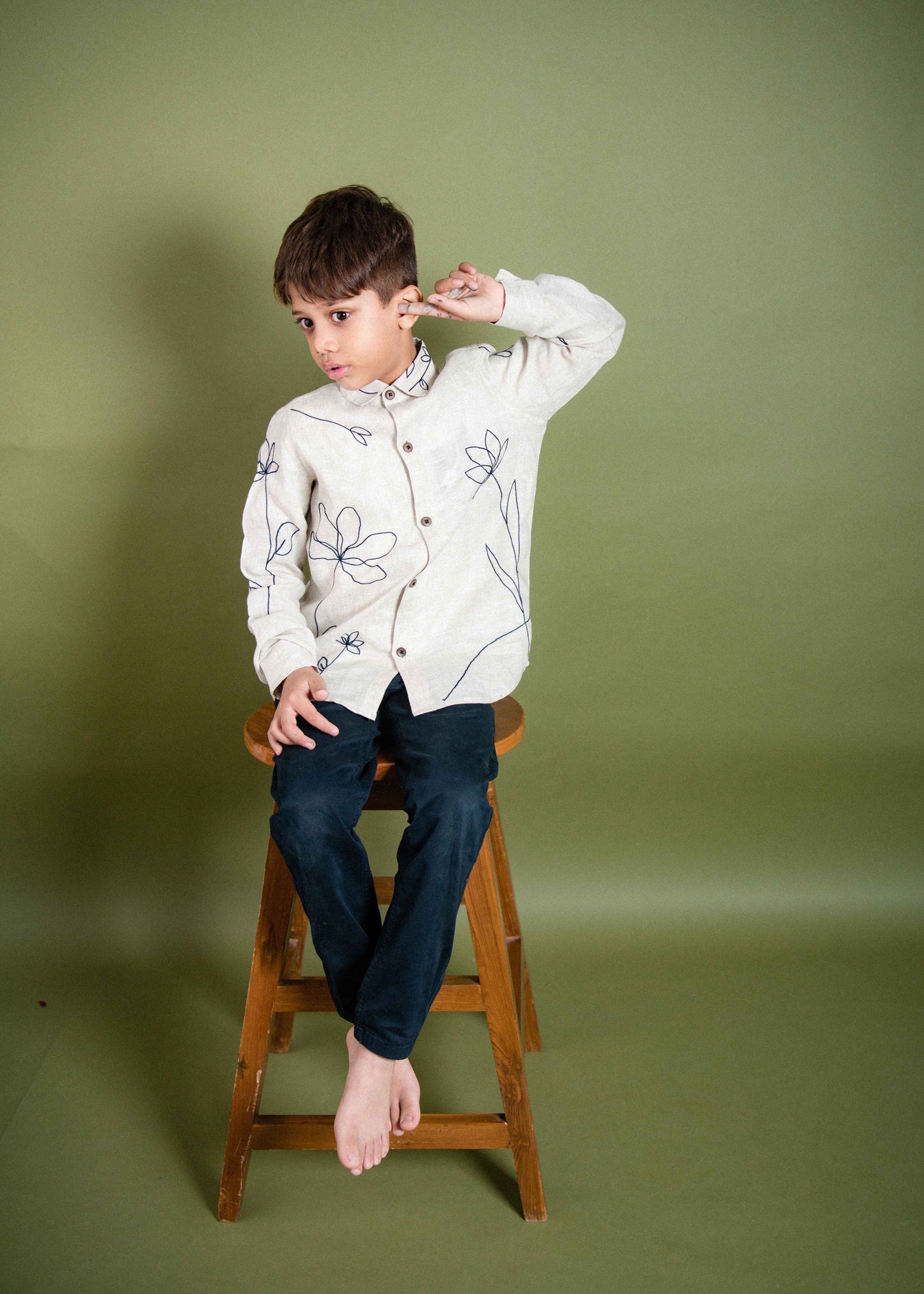 A 5 year old boy in a full sleeved floral embroidered shirt sitting on a stool