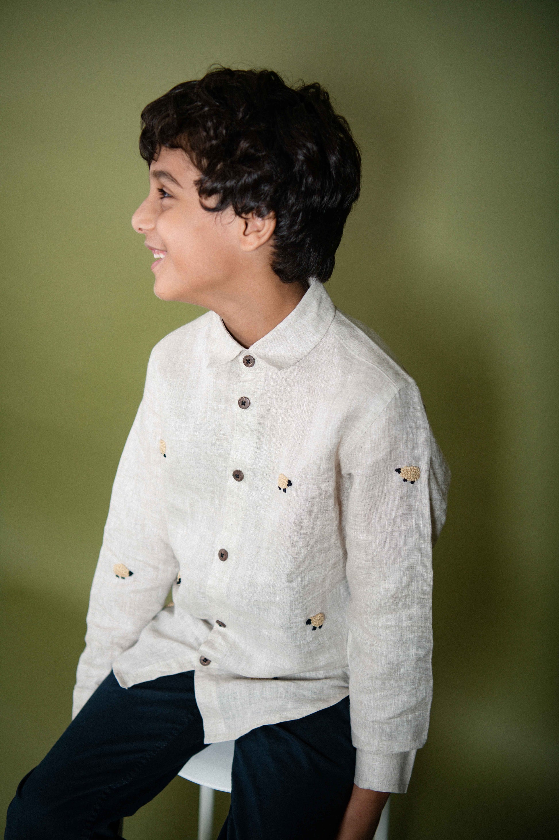 A young 7-8 year old boy in an off-white linen full sleeved shirt with small sheep embroidered throughout with his side profile visible