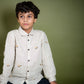 A young 7-8 year old boy in an off-white linen full sleeved shirt with small sheep embroidered throughout sitting against an olive green background