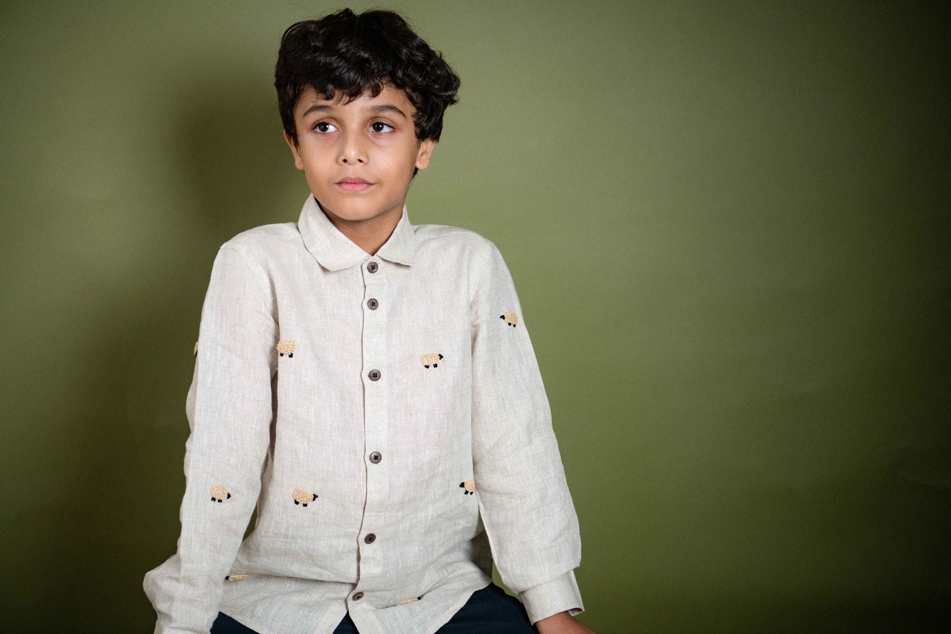 A young 7-8 year old boy in an off-white linen full sleeved shirt with small sheep embroidered throughout sitting against an olive green background