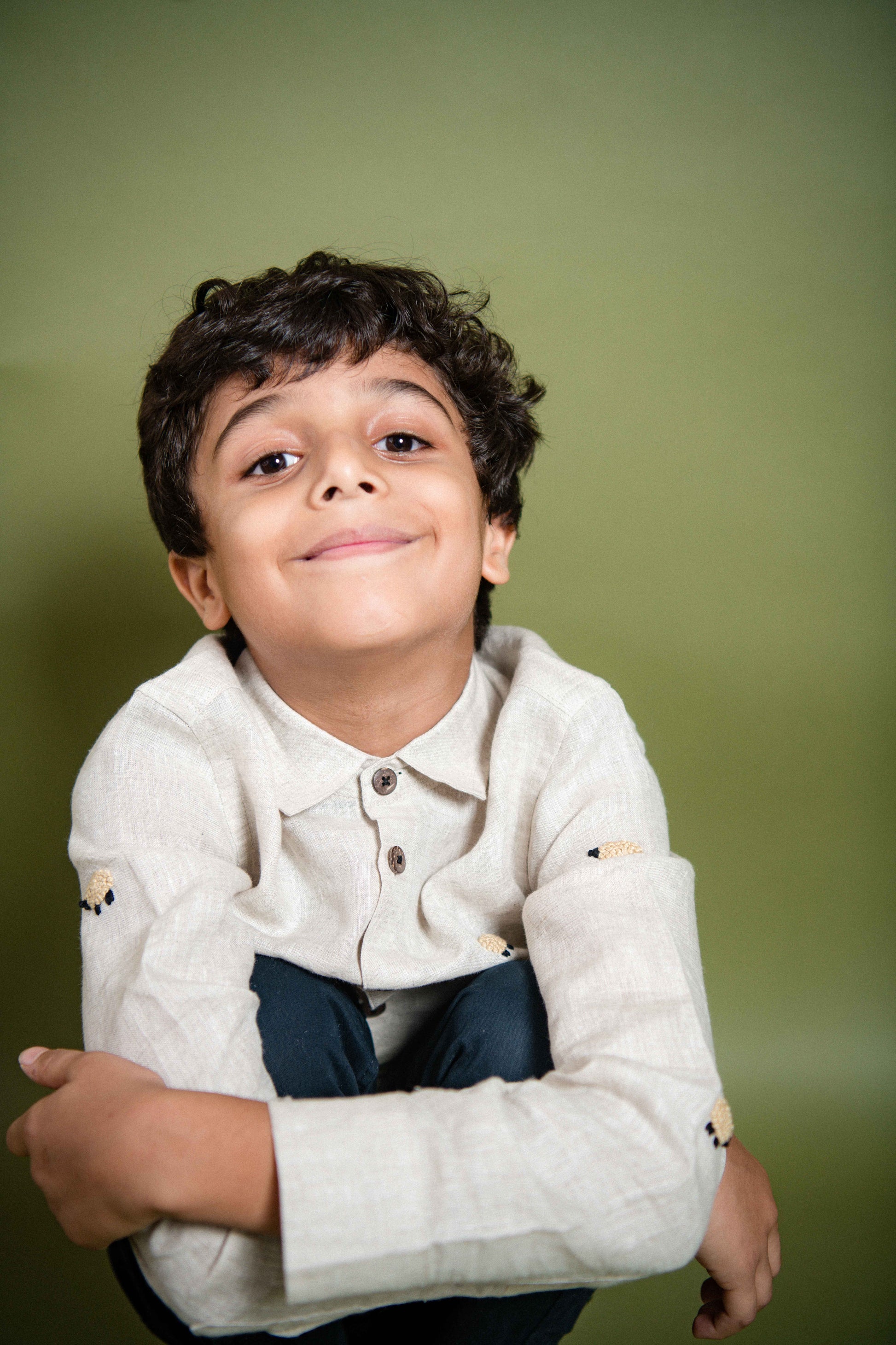 A young 7-8 year old boy in an off-white linen full sleeved shirt with small sheep embroidered throughout sitting on a chair looking cute