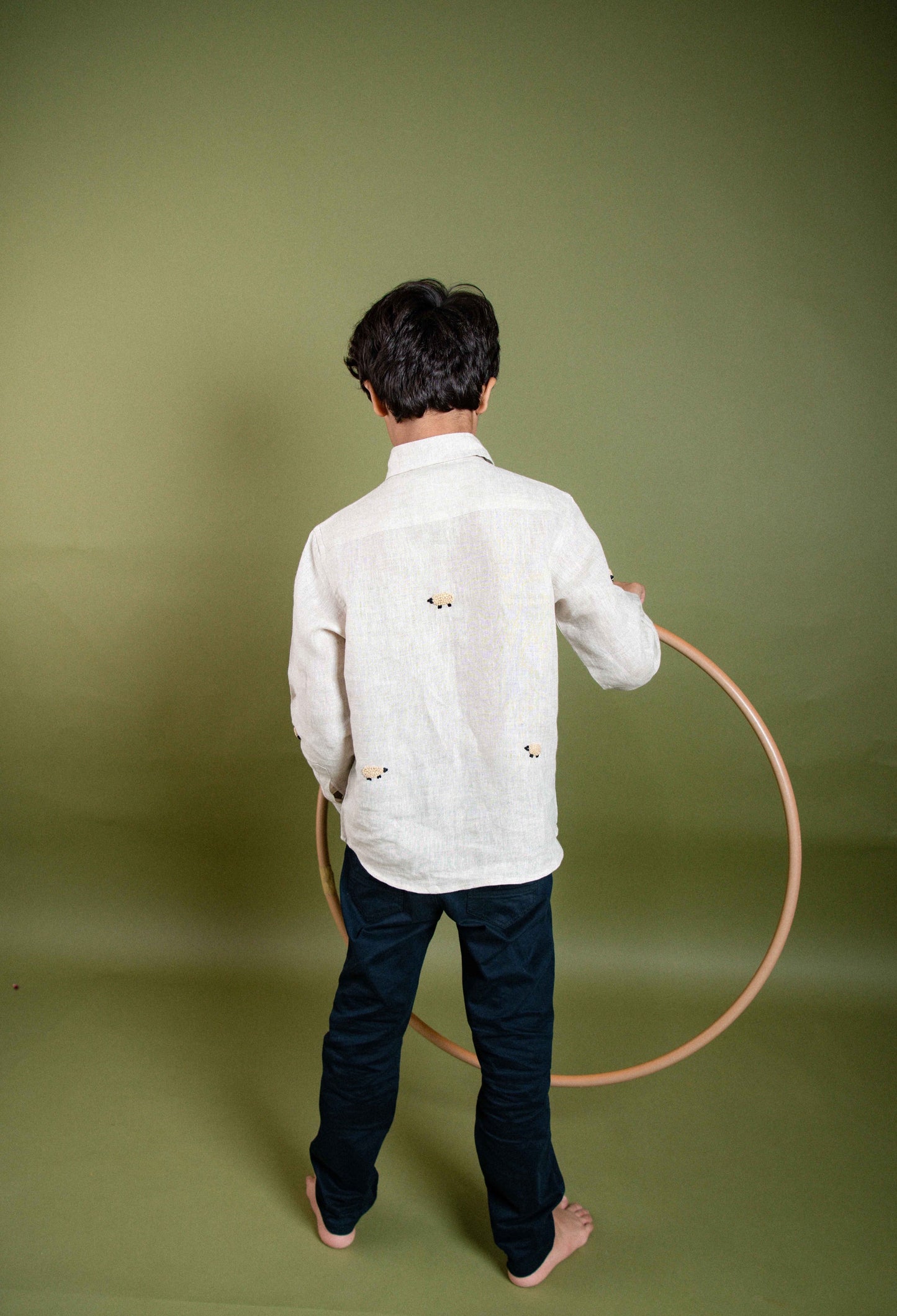 A young 7-8 year old boy in an off-white linen full sleeved shirt with small sheep embroidered throughout. He is playing with a hulahoop and the details of the back are highlighted