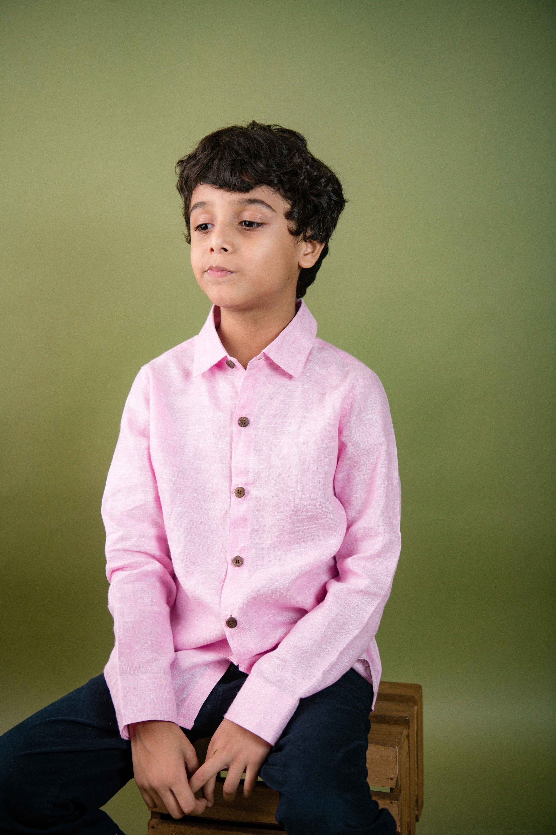 A young 7-8 year old boy in a pink Long sleeved linen shirt is sitting on a crate