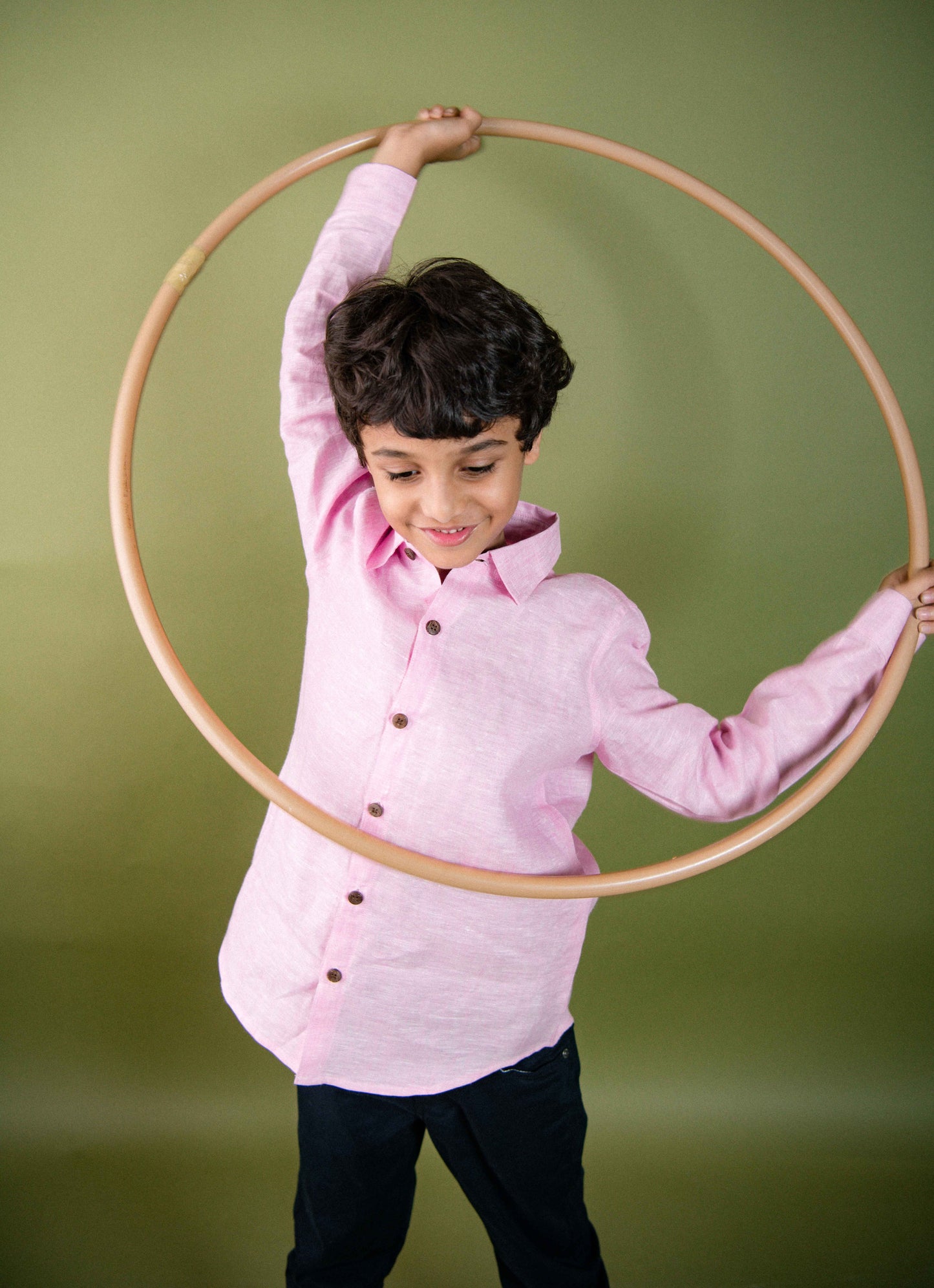 A young 7-8 year old boy in a pink Long sleeved linen shirt is playing with a hula=hoop