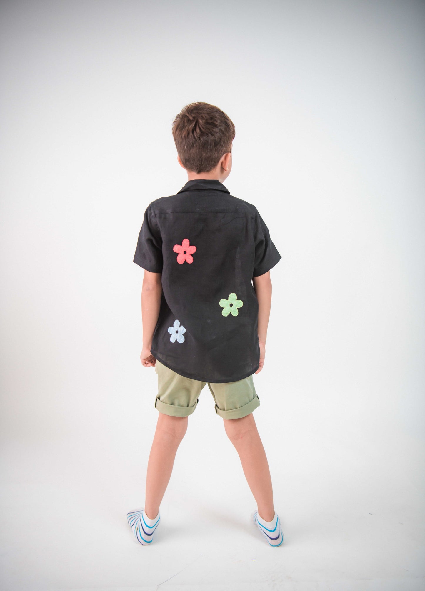 Back of a young 5-6 year old boy in a floral black linen shirt standing against a white background