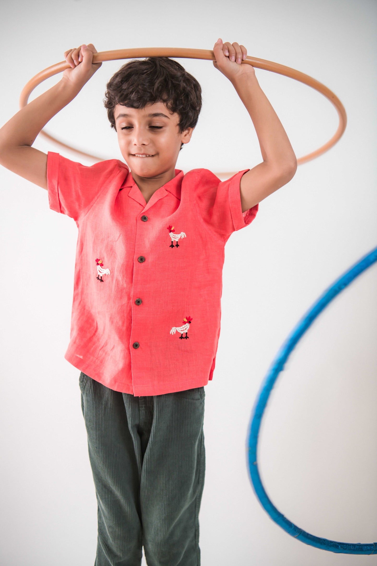 A 5-6 year old boy in a red linen shirt with embroidered roosters is playing with a hula hoop