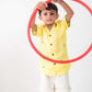 a small 4-year boy in a yellow linen shirt with small green embroidered flowers is playing with a red hula hoop