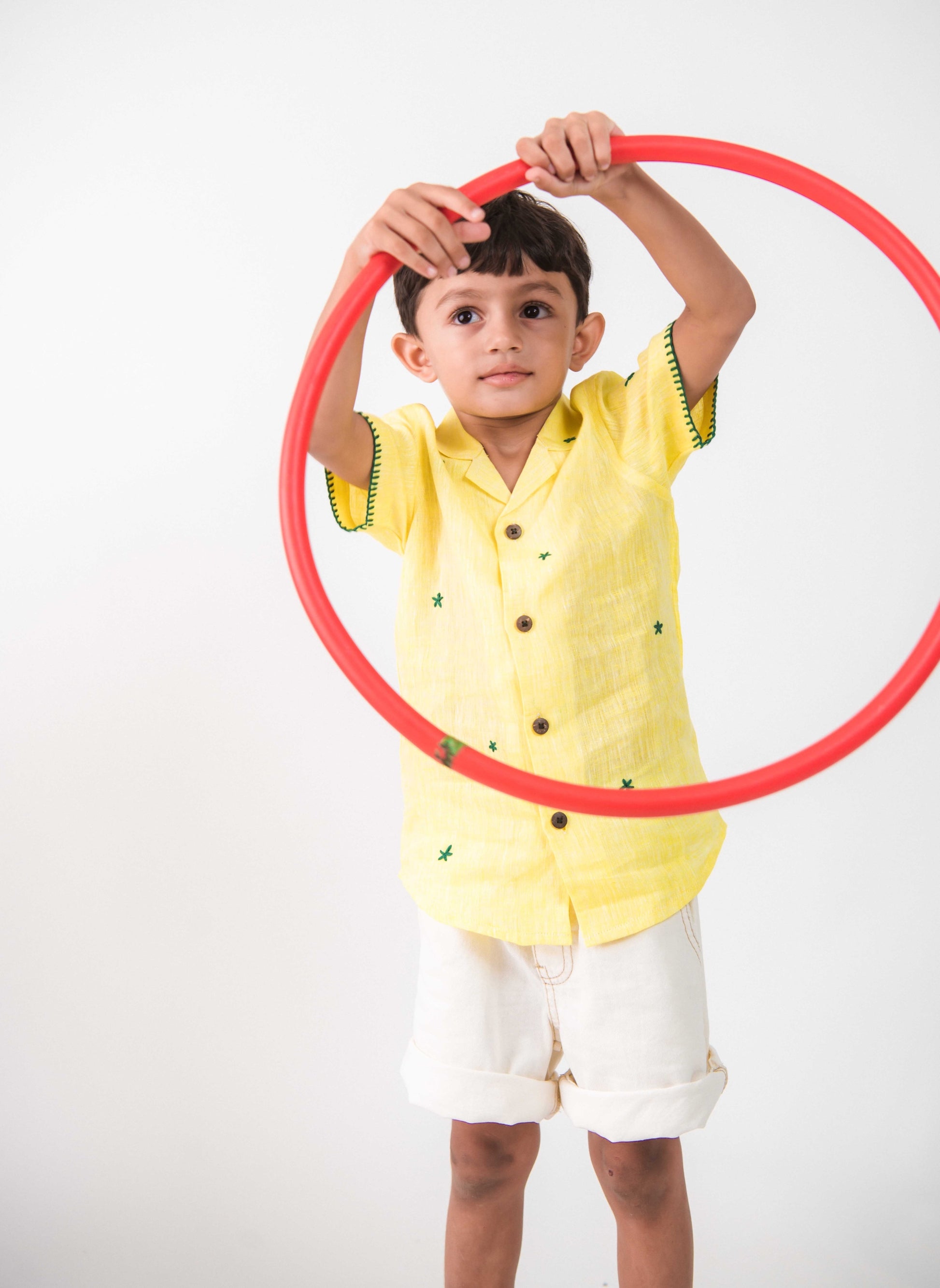 a small 4-year boy in a yellow linen shirt with small green embroidered flowers is playing with a red hula hoop