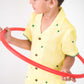 Side view of a small 4-year boy in a yellow linen shirt with small green embroidered flowers is playing with a red hula hoop