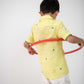 Back view of a small 4-year boy in a yellow linen shirt with small green embroidered flowers is playing with a red hula hoop