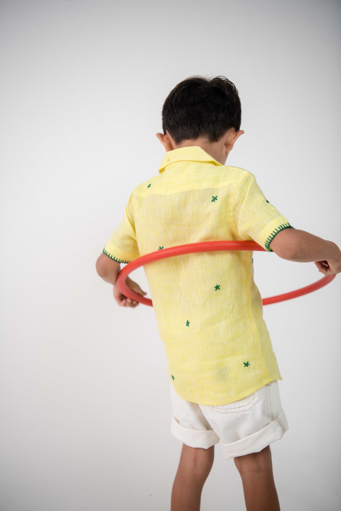 Back view of a small 4-year boy in a yellow linen shirt with small green embroidered flowers is playing with a red hula hoop