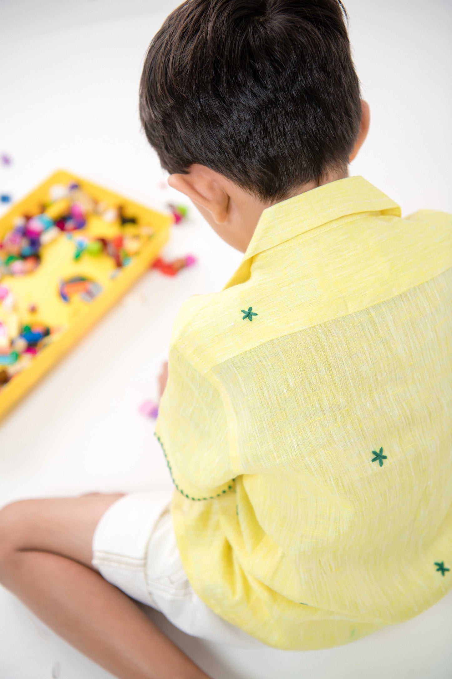 a small 4-year boy in a yellow linen shirt with small green embroidered flowers is sitting and playing with legos