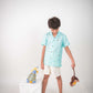 A young 7-8 year old boy in a blue linen half sleeved shirt with small sheep embroidered throughout is standing against a white background playing with toys