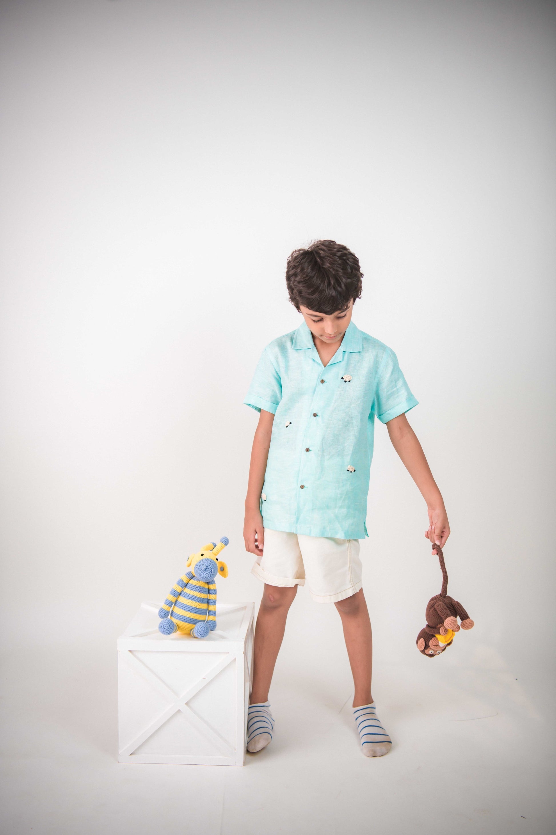 A young 7-8 year old boy in a blue linen half sleeved shirt with small sheep embroidered throughout is standing against a white background playing with toys