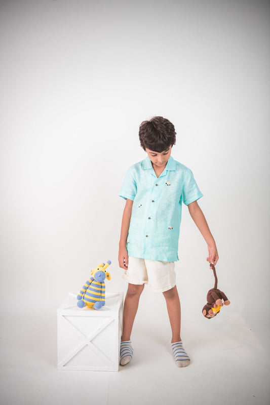 A young 7-8 year old boy in a blue linen half sleeved shirt with small sheep embroidered throughout is standing against a white background playing with toys