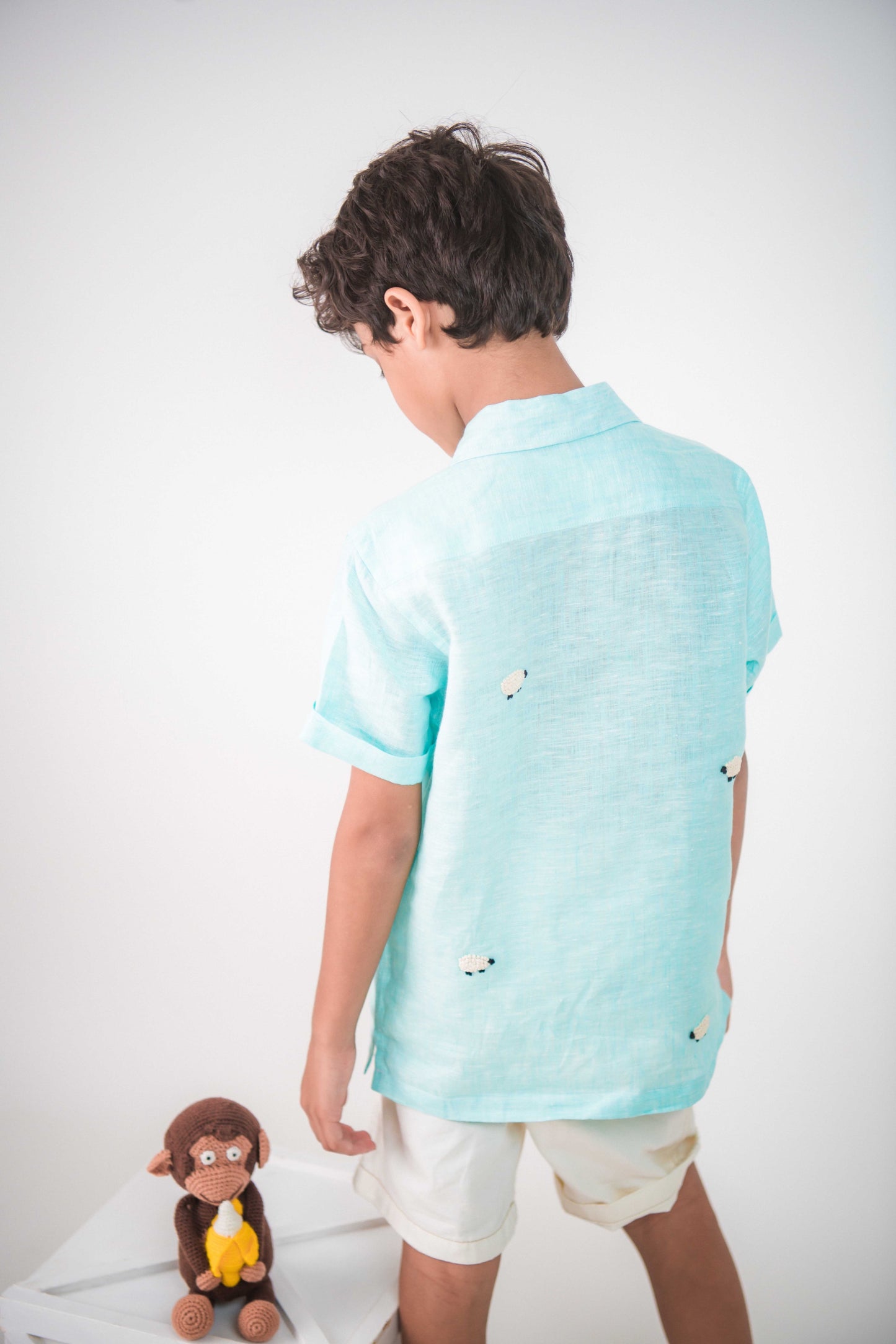 Back details of a young 7-8 year old boy in a blue linen half sleeved shirt with small sheep embroidered throughout is standing against a white background