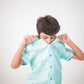 A young 7-8 year old boy in a blue linen half sleeved shirt with small sheep embroidered throughout is sitting against a white background