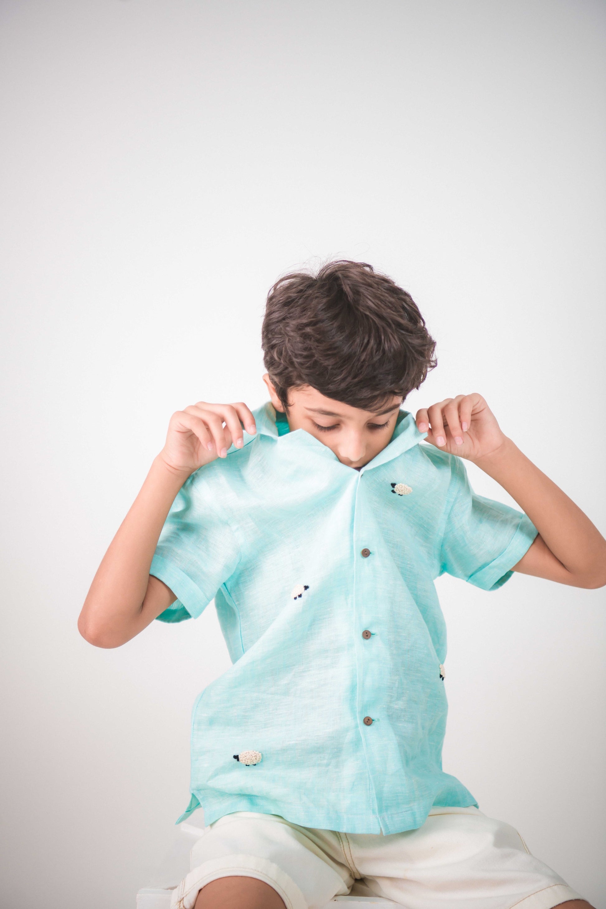 A young 7-8 year old boy in a blue linen half sleeved shirt with small sheep embroidered throughout is sitting against a white background