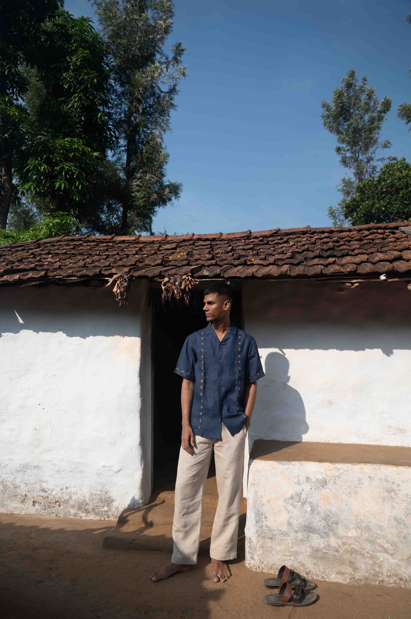 A man in eco-friendly, buds in blue hand-embroidered linen shirt in front of a hut. half sleeves shirt, Material: linen, front view