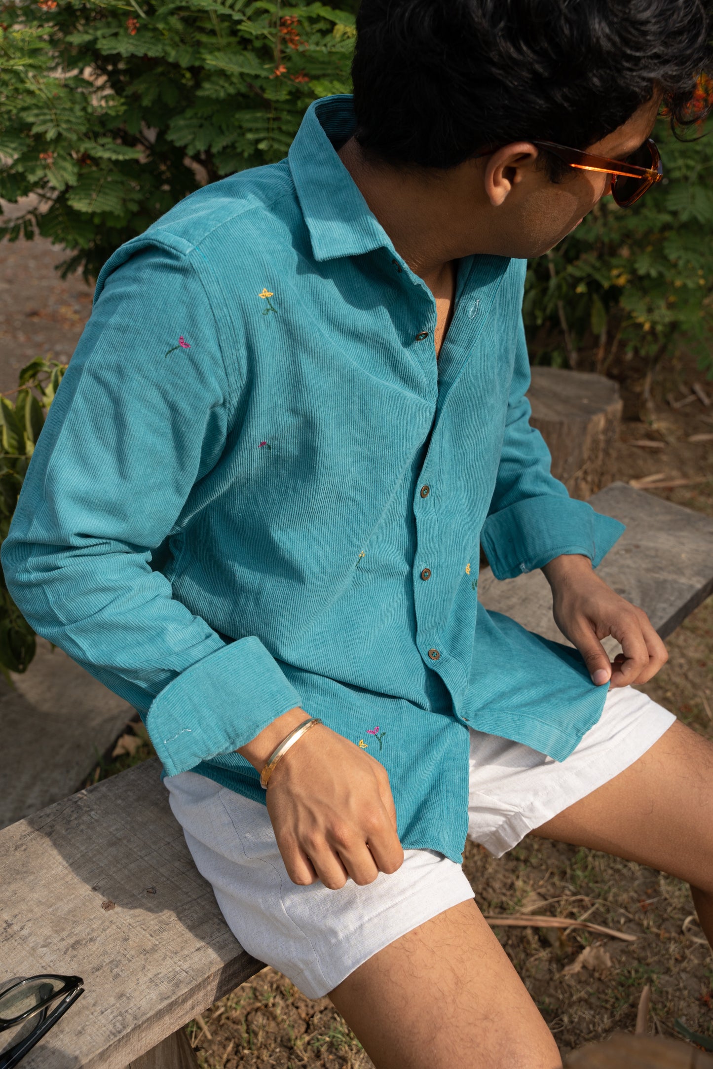 Pranav in teal cotton corduroy teal overshirt with small hand-embroidered flowers is standing in front of trees 