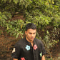 A young man in a black linen shirt with colorful flowers coming out of a grove of trees 